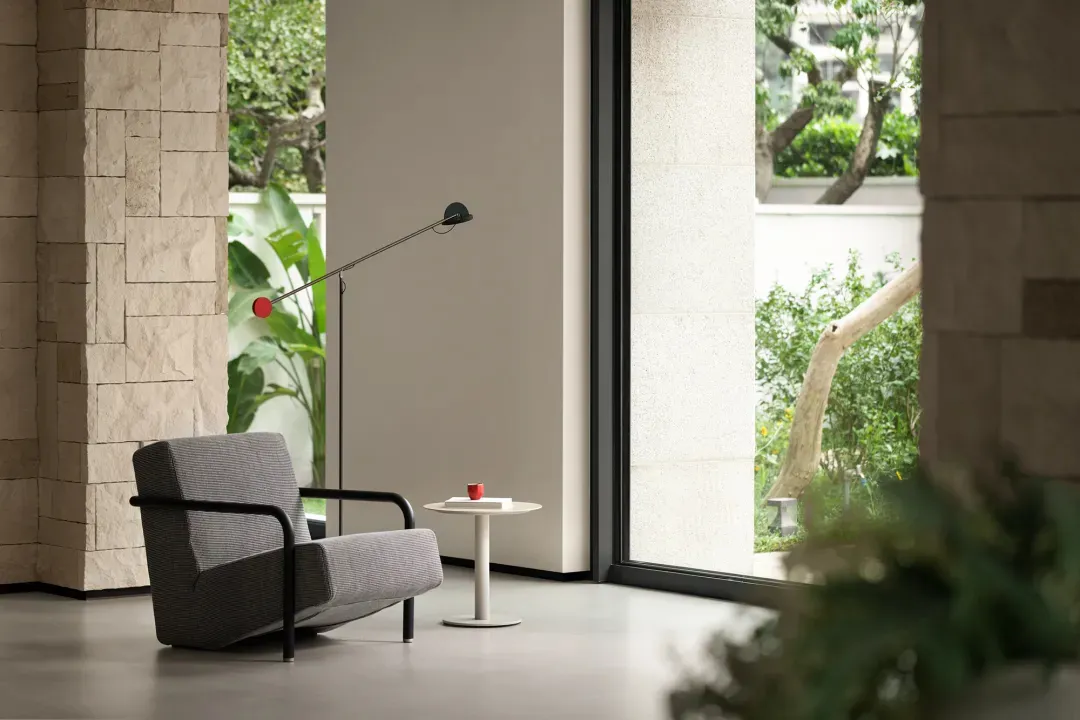 A gray chair sits in front of a window in a calm and inviting living space with natural light and two potted plants nearby