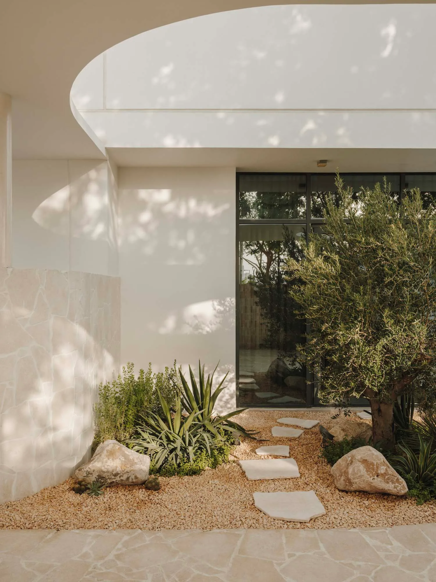 A serene outdoor patio with a rock pathway, potted plants, and two benches near a doorway, surrounded by a coastal villa