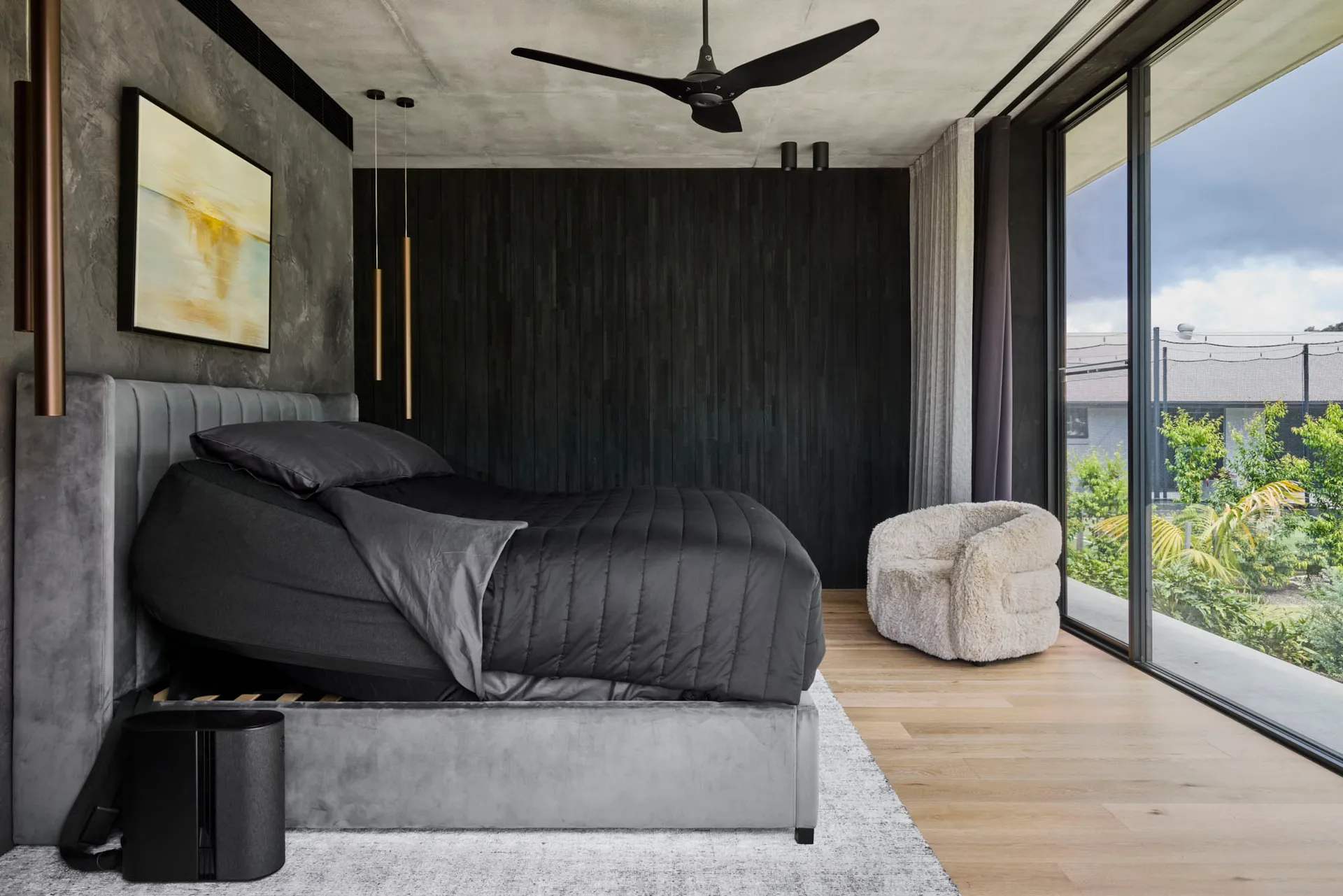 A modern bedroom with a black bed on a wooden floor, gray and white tones, ceiling fans, a chair, and a potted plant