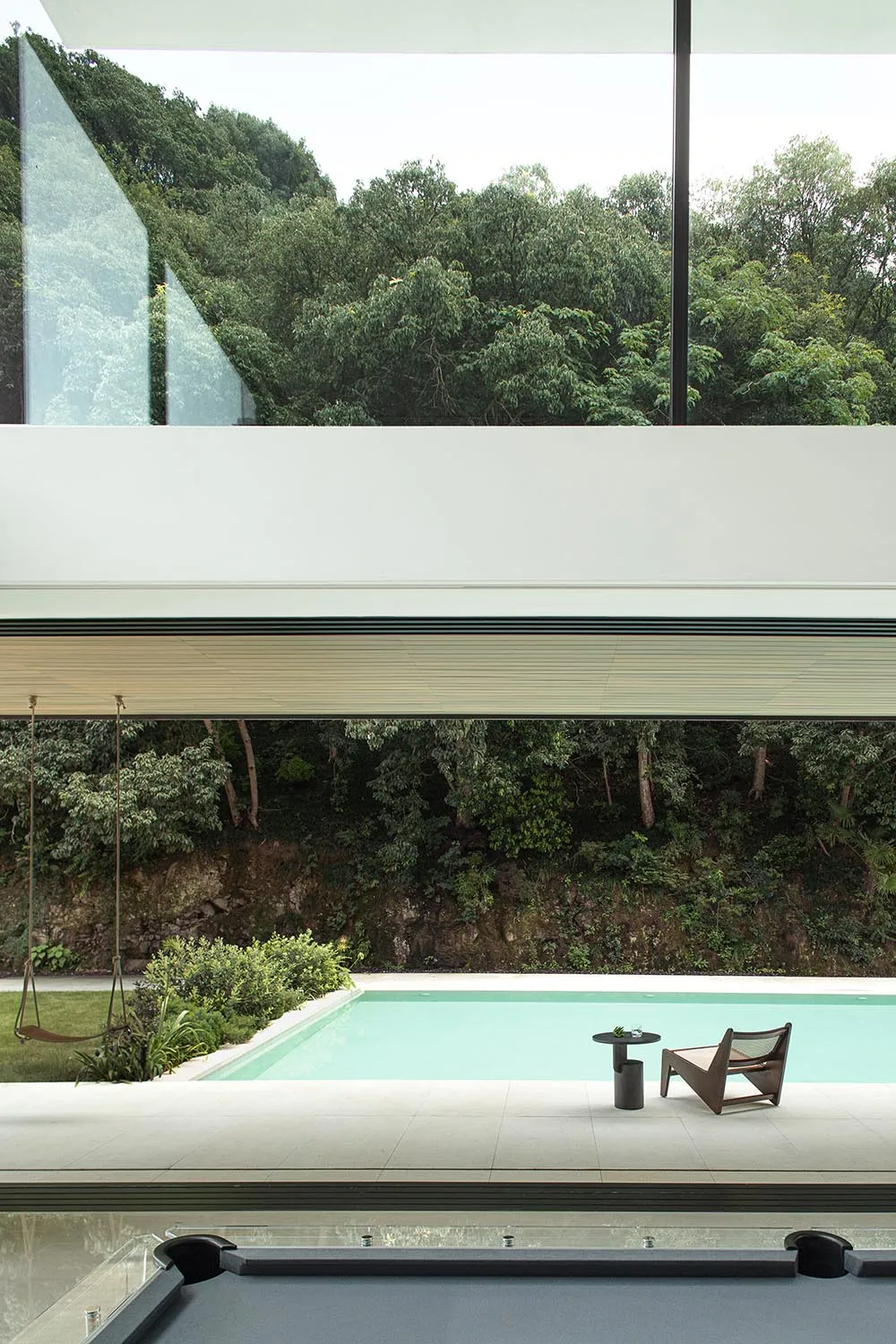Indoor pool area with a large glass wall offering a view of the surrounding forest, two chairs, and an empty pool