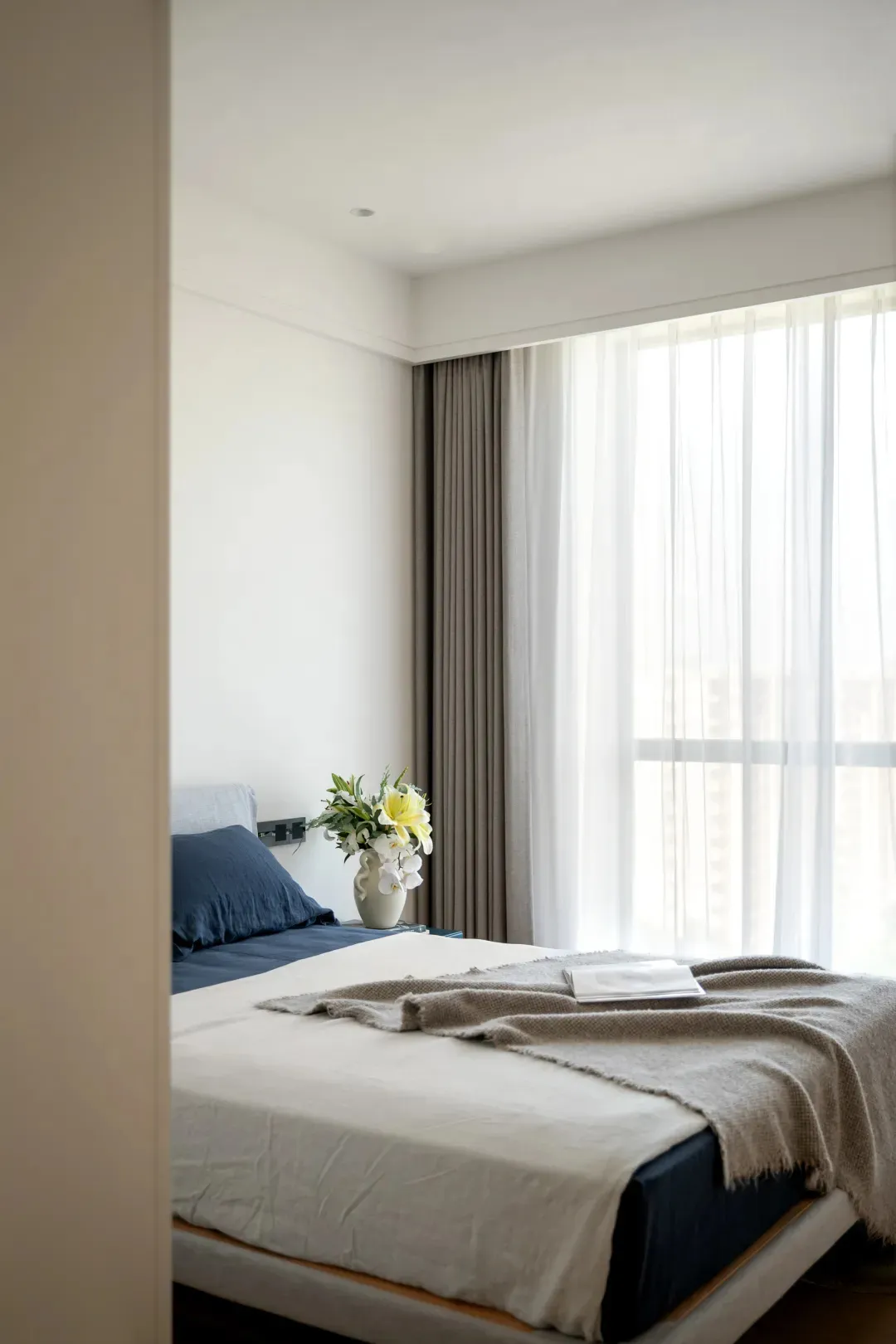 A serene bedroom with a white bed, two vases, and a book, surrounded by natural light from a window