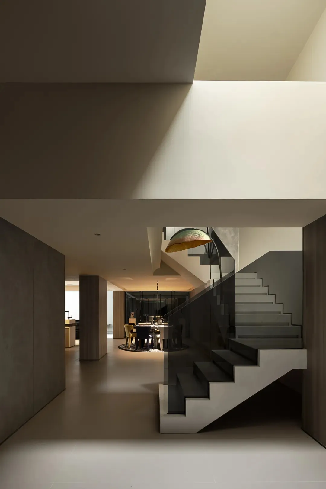 A modern staircase with concrete steps and a glass railing leads to a dining area with a table and chairs, and a couch nearby