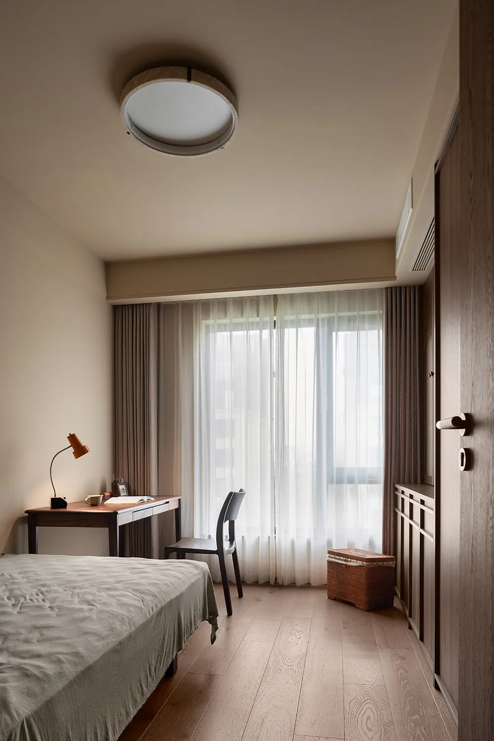 A bedroom with a bed and desk, natural light from two windows, and a lamp, set against a clean and bright white wall background