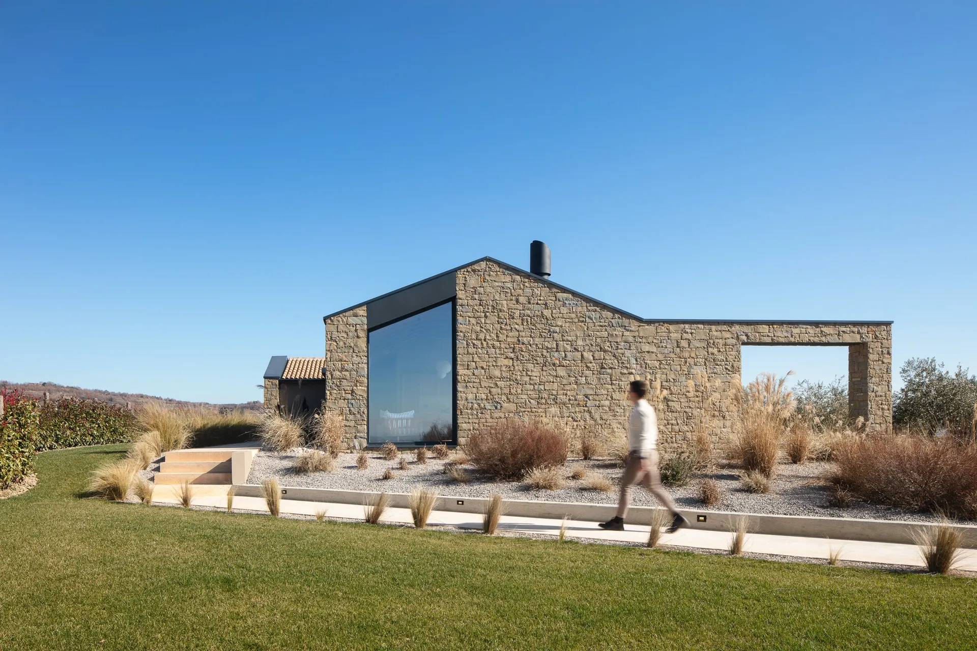 A stone house with large windows, potted plants, and a man walking in front of it