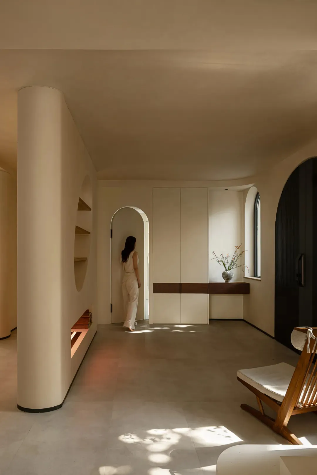 A cozy living area with a woman standing in a hallway between two rooms, featuring two chairs and a potted plant, creating a warm and inviting atmosphere