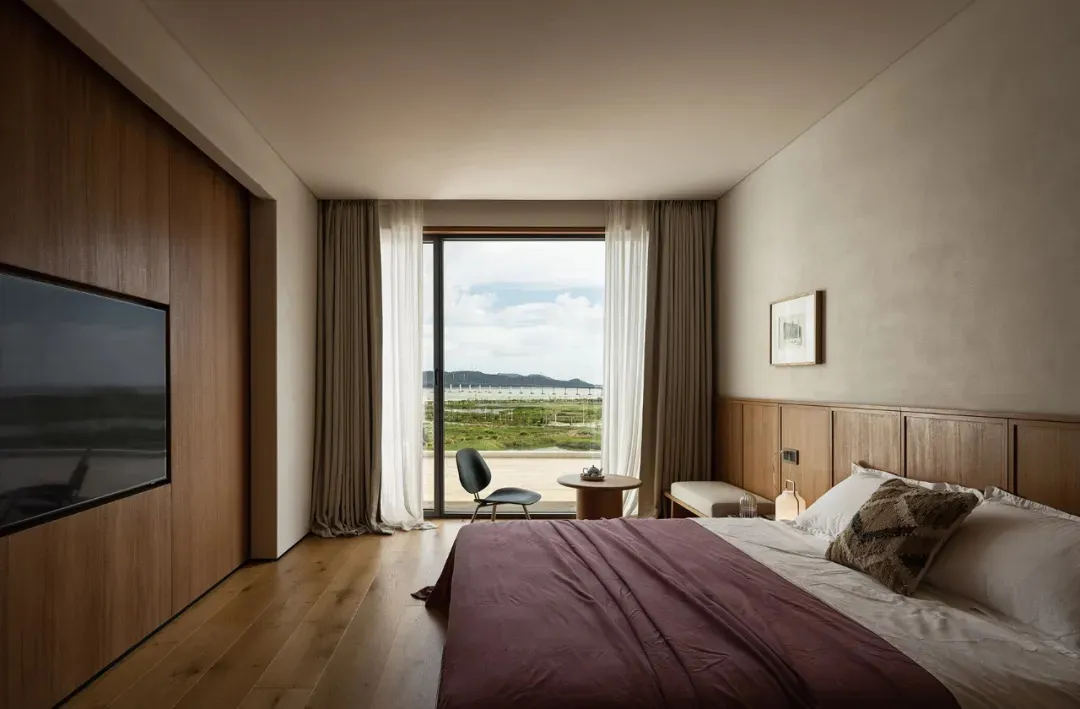 A serene bedroom with two beds, a chair, and a dining table, offering a view of the ocean through a large window