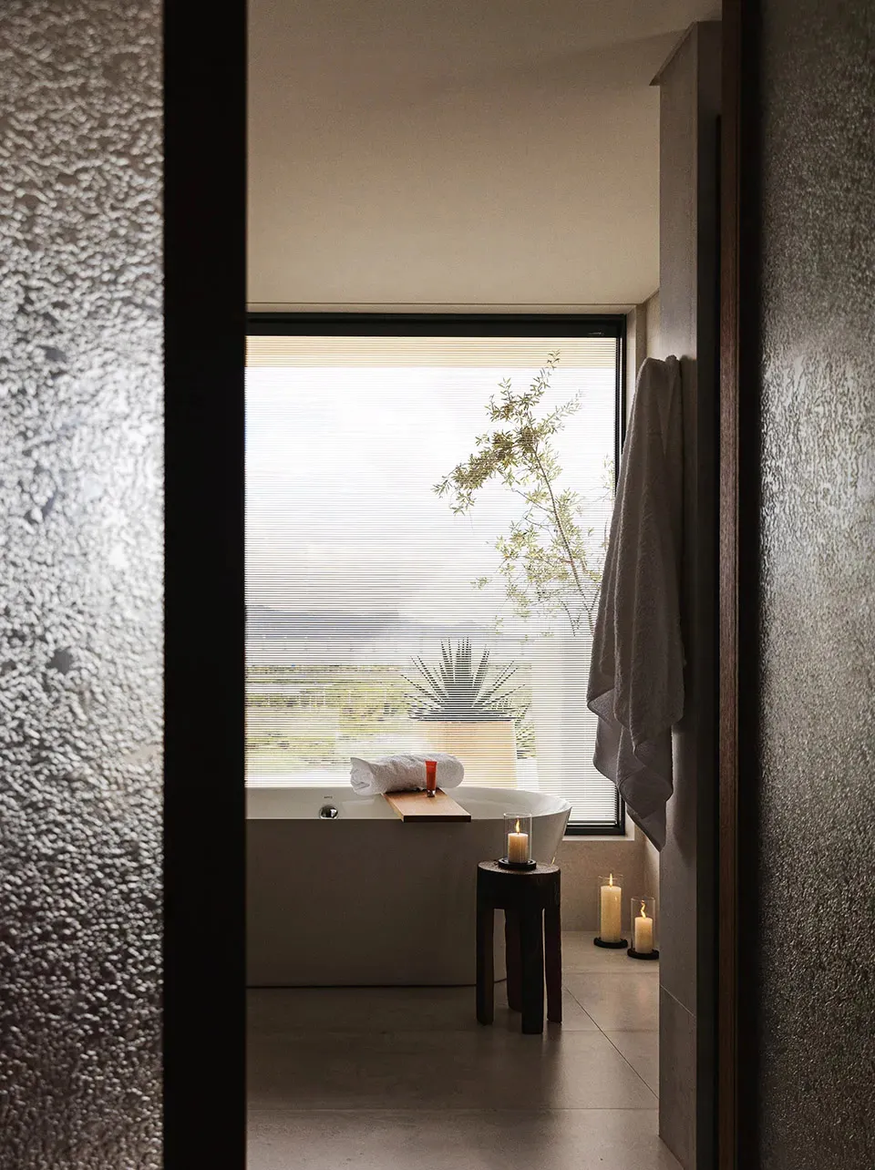 A modern bathroom with a bathtub next to a window, candles, and a potted plant, creating a bright and inviting atmosphere