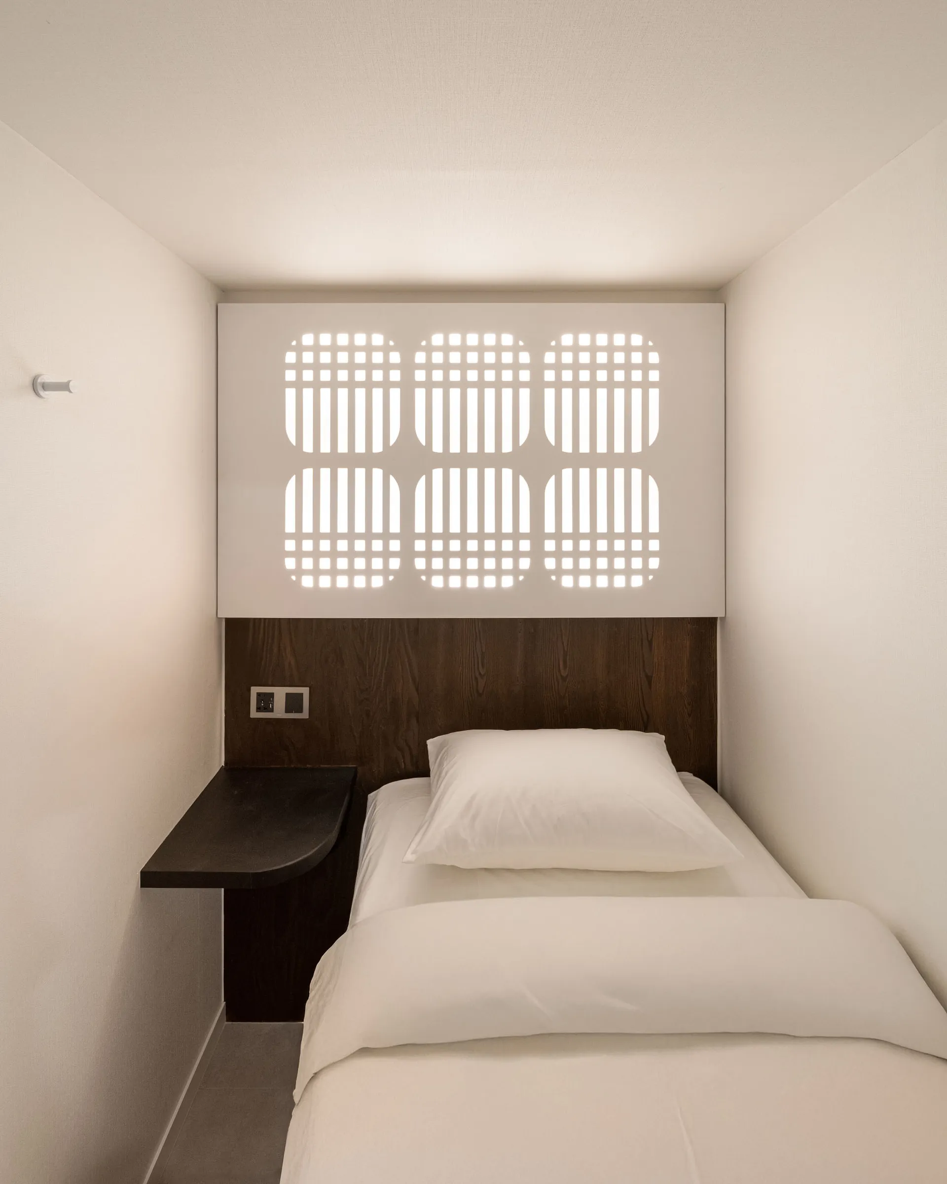 A modern bedroom with a white comforter and brown headboard, a desk, and a chair, well-lit by natural light through a window