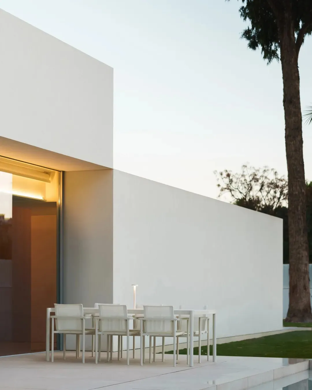 A modern outdoor patio with a dining table and white chairs, surrounded by a white building and a potted plant, creating a comfortable and inviting atmosphere