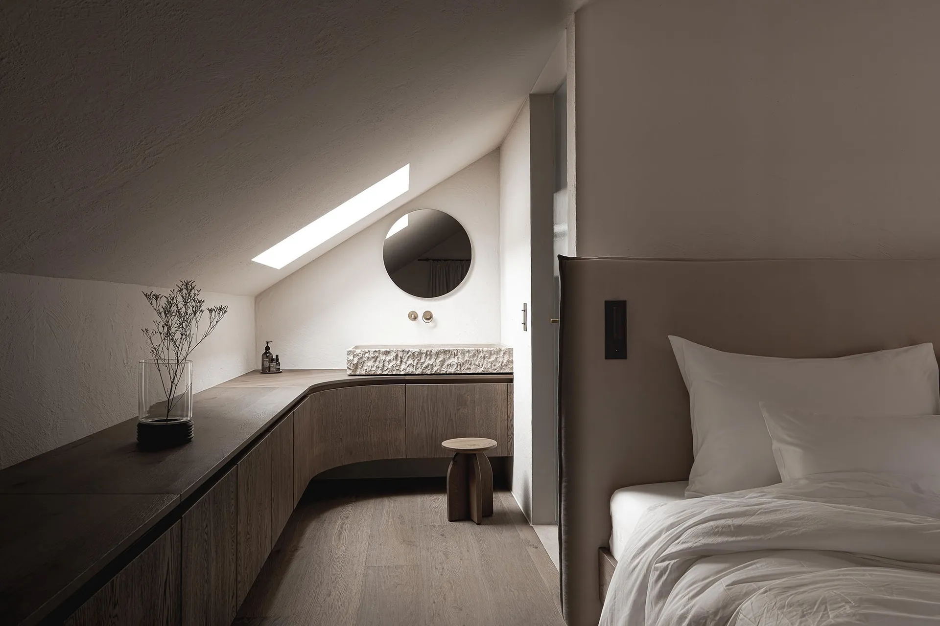 Dimly lit bedroom with a bed, sink, and mirror, featuring wood materials and neutral colors