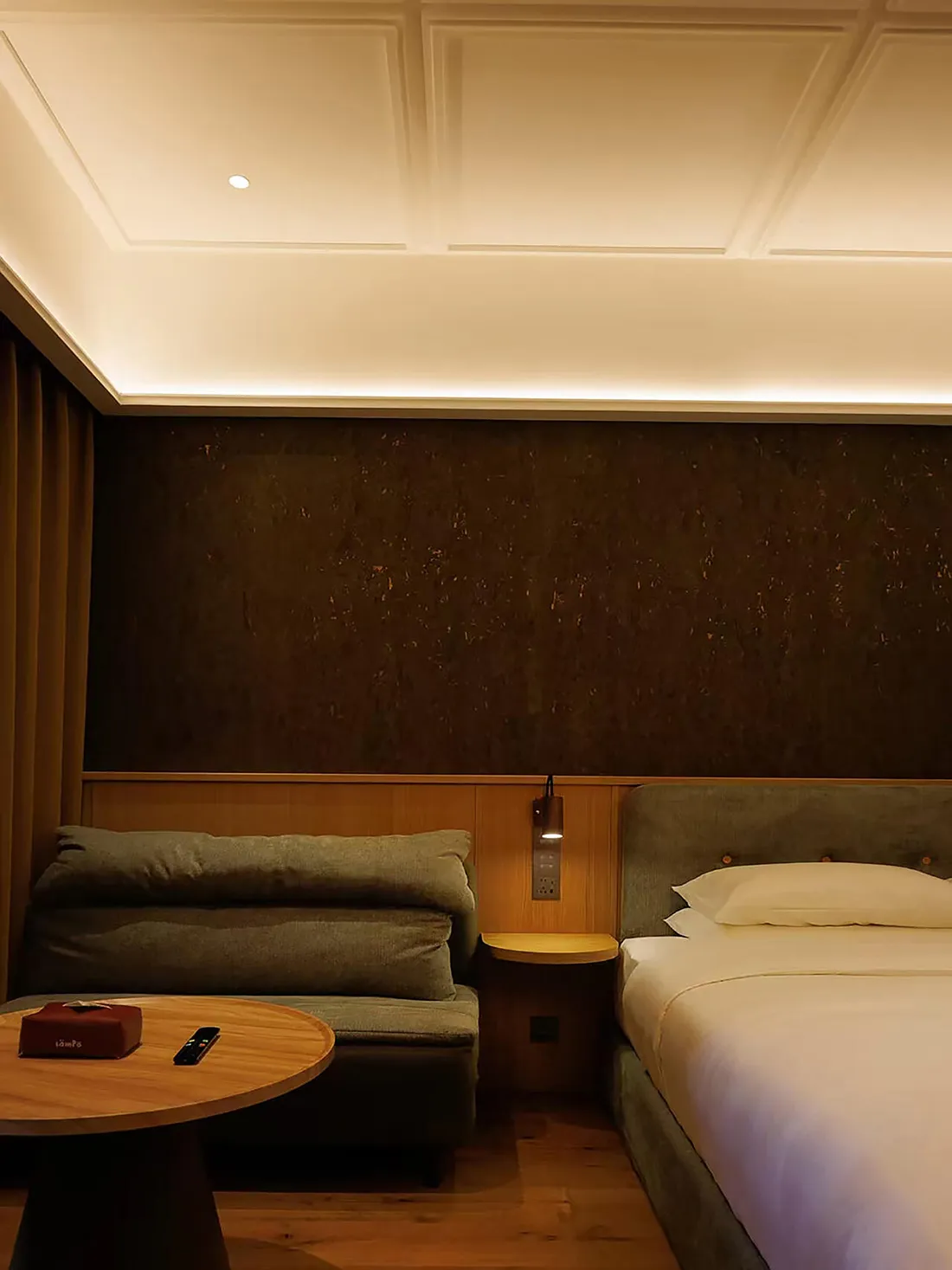 Cozy bedroom with a wooden floor, a bed on the right, a couch on the left, and a table in the middle, decorated with brown and white colors in a dimly lit space