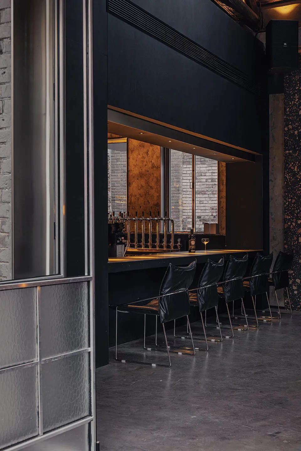 A modern bar with a black counter, wine glass, and bottles, surrounded by chairs in a well-lit restaurant or bar setting