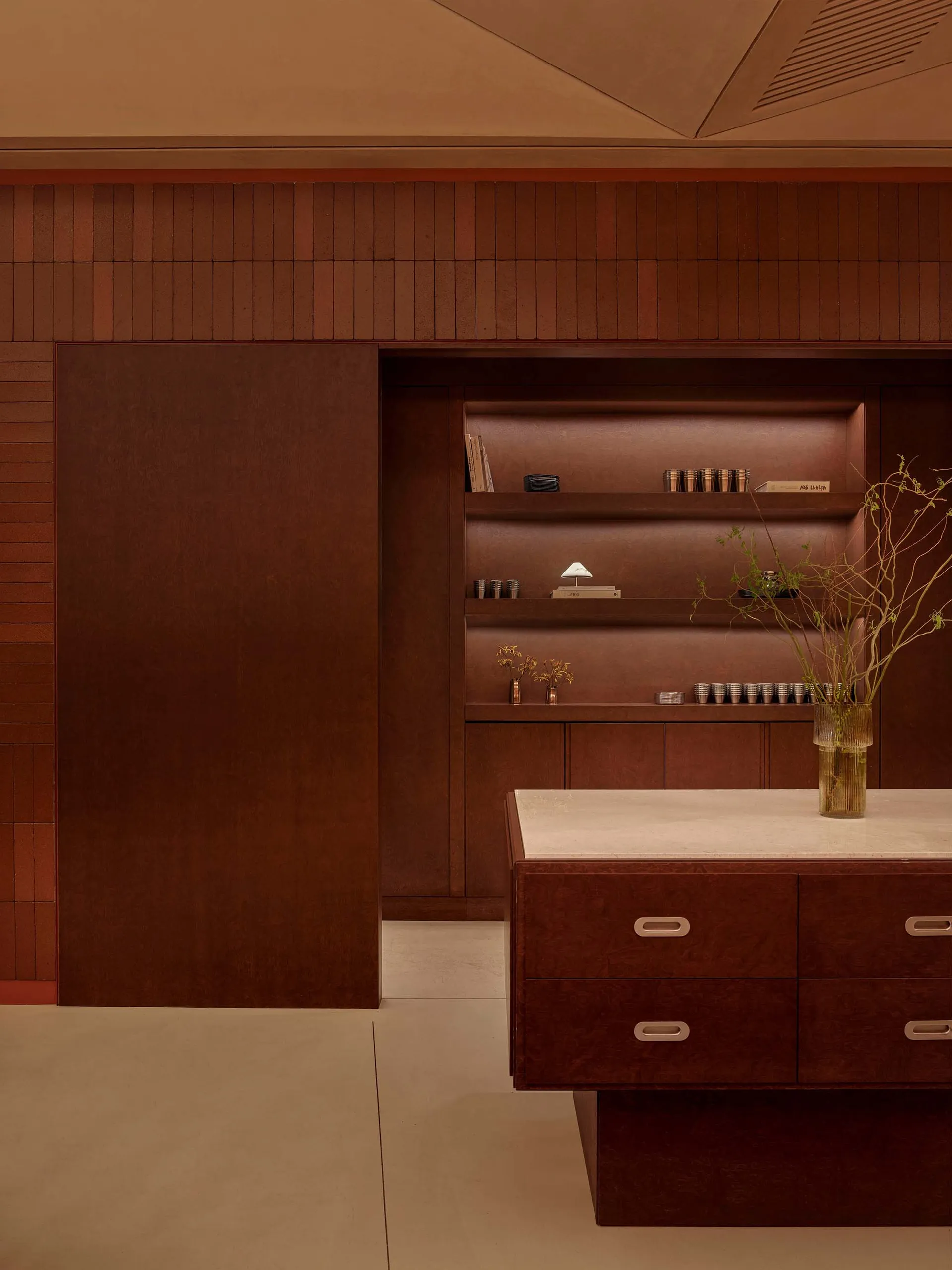 Bathroom with a wooden cabinet, marble counter, and a vase of flowers, set against a brown color scheme with a potted plant and dim lighting