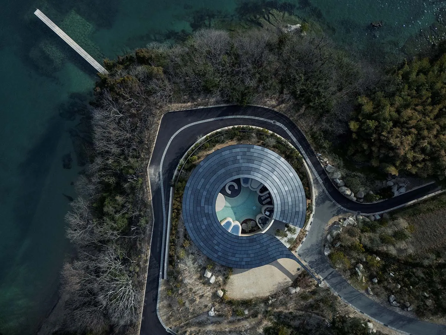 Photograph of a modern house with a circular pool house surrounded by trees and situated near a body of water