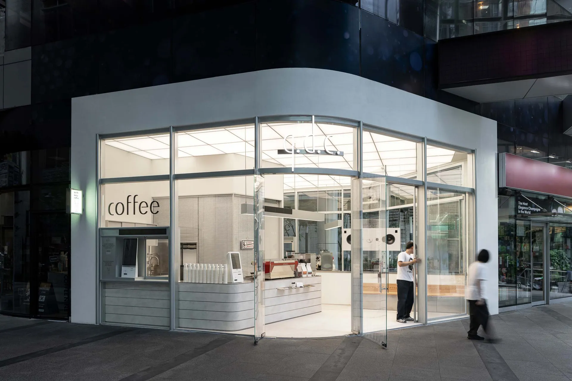 A modern coffee shop with a glass front, white interior, and natural light, featuring appliances, furniture, and a welcoming atmosphere