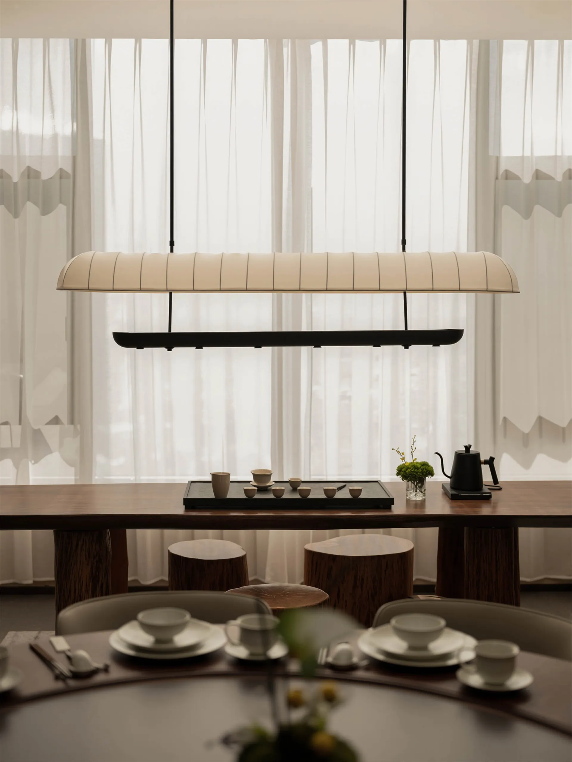 Dining room with a wooden table, chairs, and a potted plant, set with various items and a vase with flowers, in a warm and inviting space with natural lighting