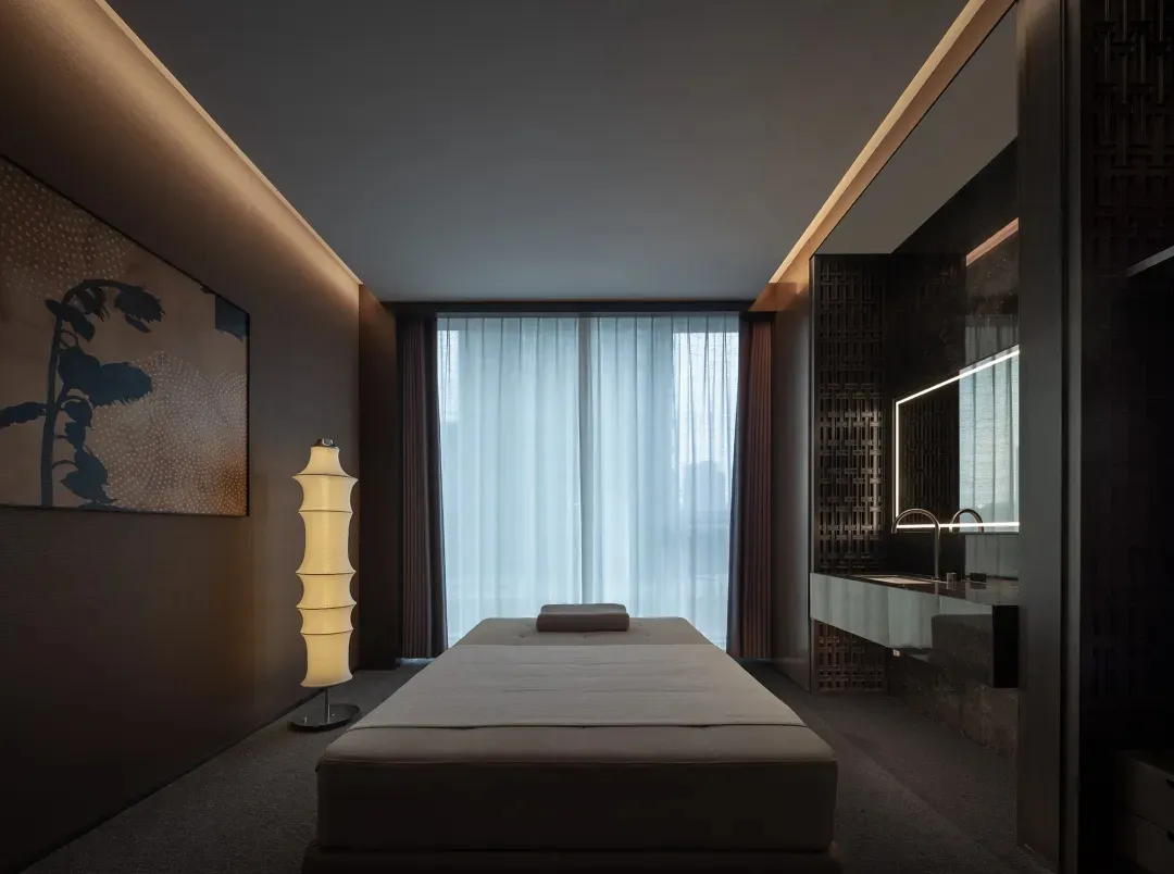 A dimly lit bedroom with a white bed, a wooden bed frame, and a brown window frame, set against a neutral background