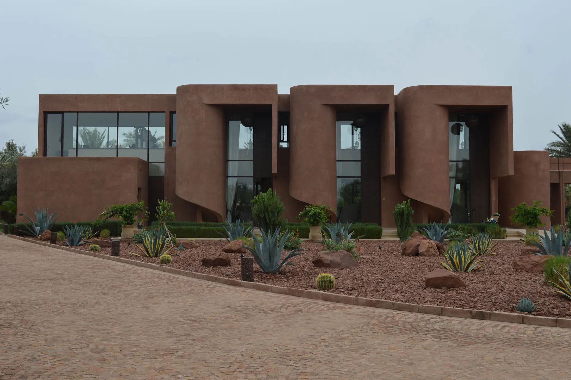 A modern building with a unique design, surrounded by a lush courtyard with cacti and palm trees, featuring red and brown colors and a curved structure