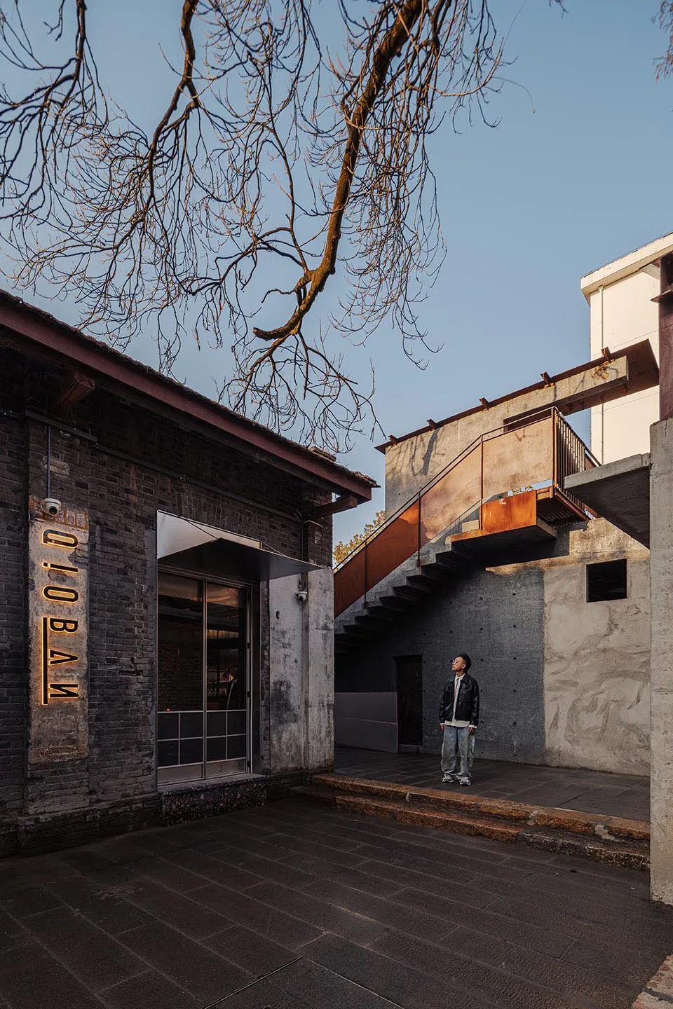 A stone building with a concrete staircase leading to a second floor, set in a quiet outdoor environment with a tree in the background