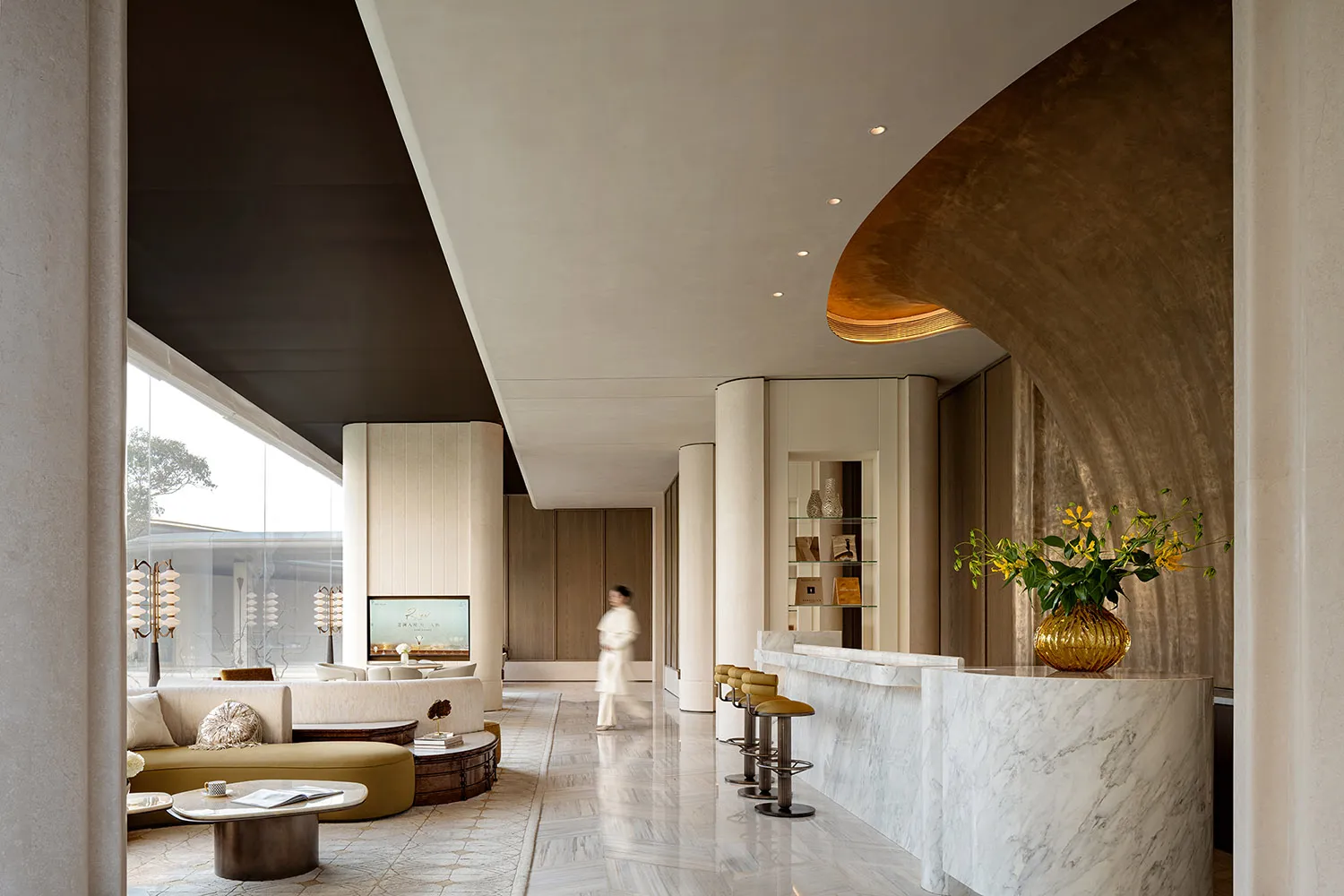 Well-lit hotel lobby with a modern design, featuring a large counter, couch, and dining table, with a woman walking through, and natural light streaming in through the windows