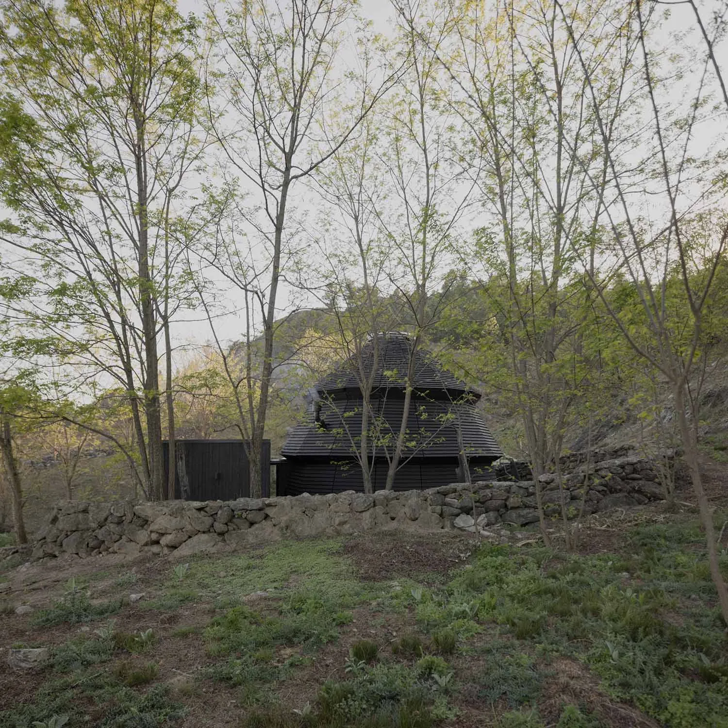 A small house or shed with a stone wall and black roof, set in a grassy area surrounded by trees