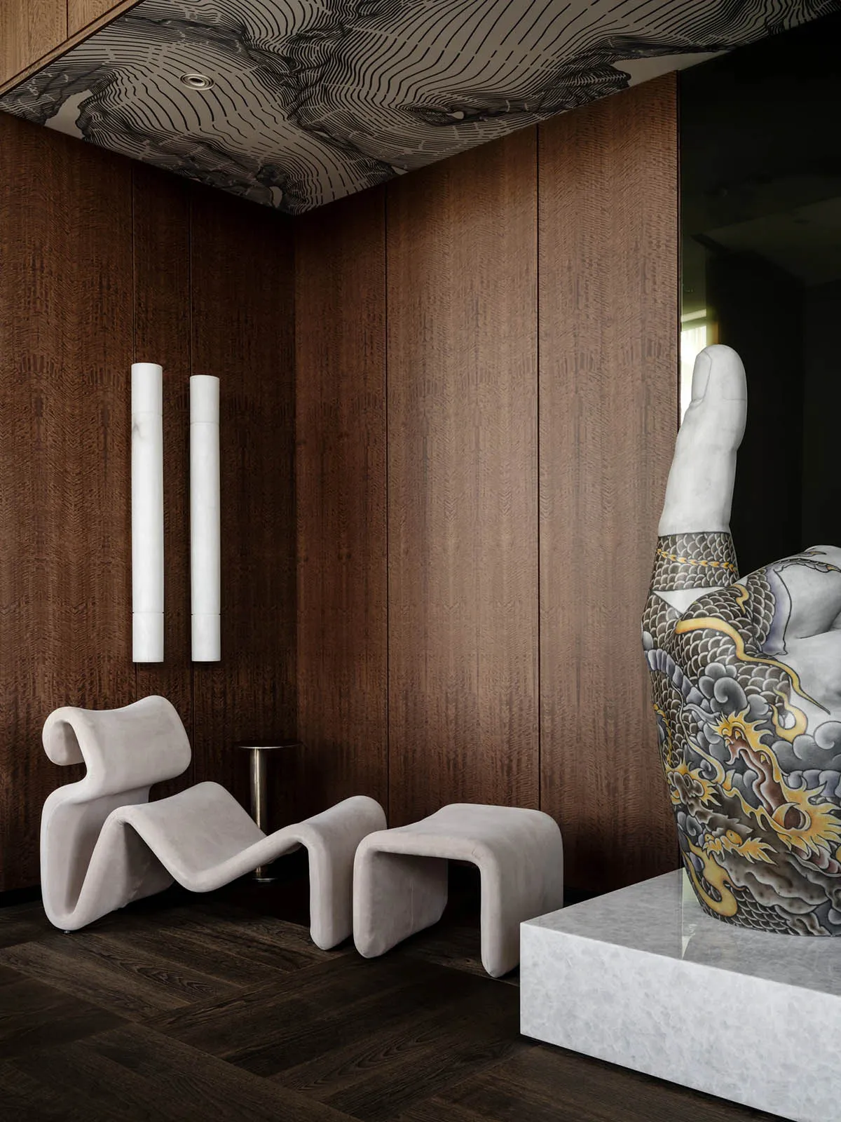 A modern room with a wooden wall, a white floor, and a large statue of a hand as the main focal point, accompanied by two chairs, a couch, and a vase, in a dimly lit space