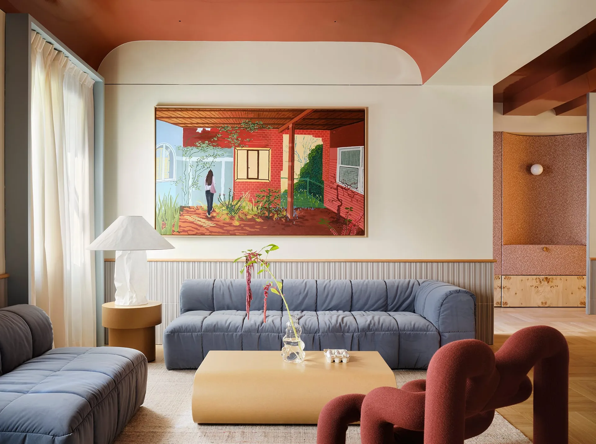 A well-lit living room with a blue couch, a chair, a dining table, potted plants, a painting, a vase, and a bowl, creating a comfortable and inviting atmosphere