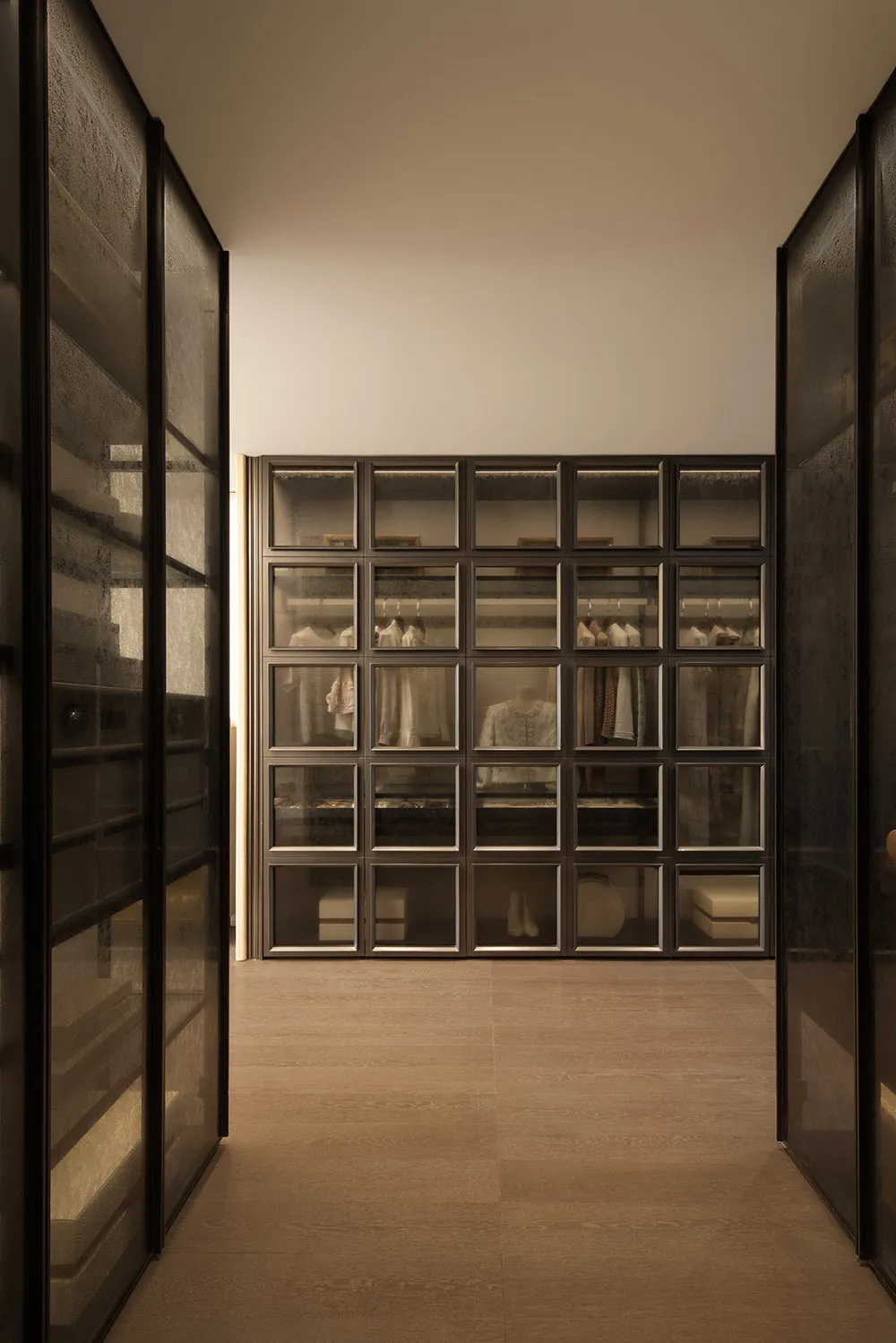 A dimly lit hallway with a wooden floor and white walls, lined with open glass doors revealing a closet with clothes and a cozy atmosphere