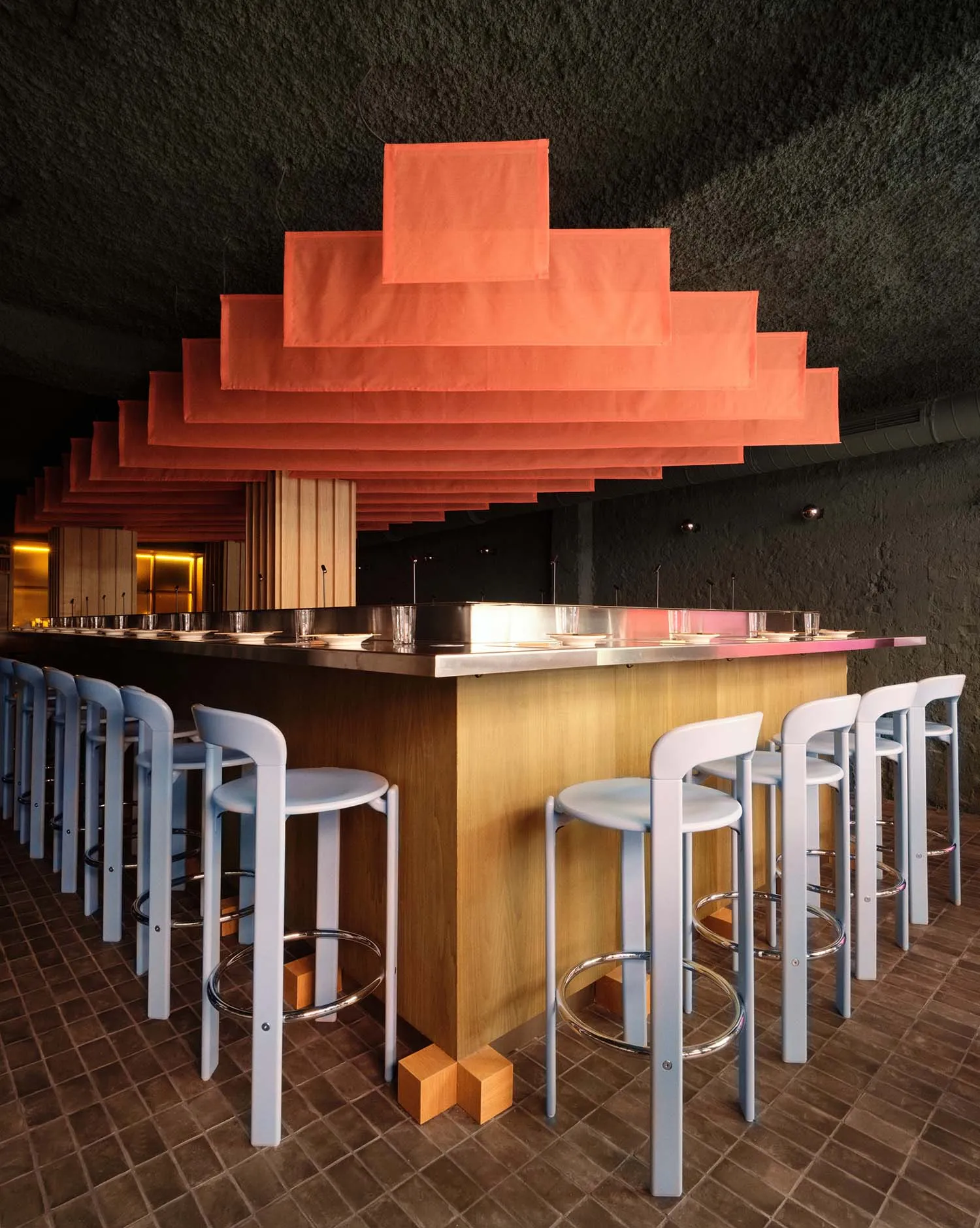 A modern bar with a wooden counter and blue stools, dim lighting, and various items on the counter