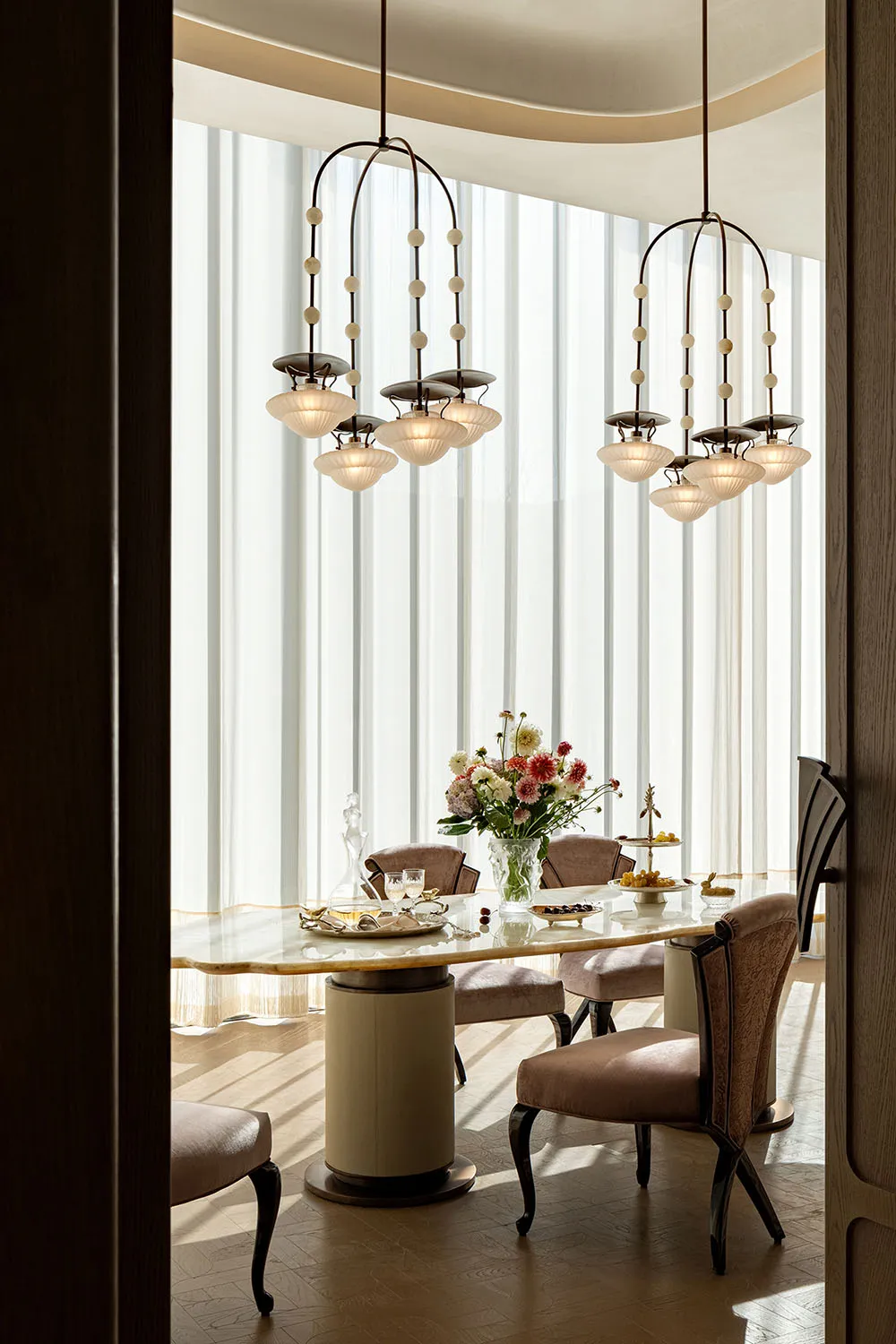 Dining room with a glass table, wood chairs, and a couch in the background, set with a vase of flowers, wine glasses, and a bowl, under soft and warm lighting
