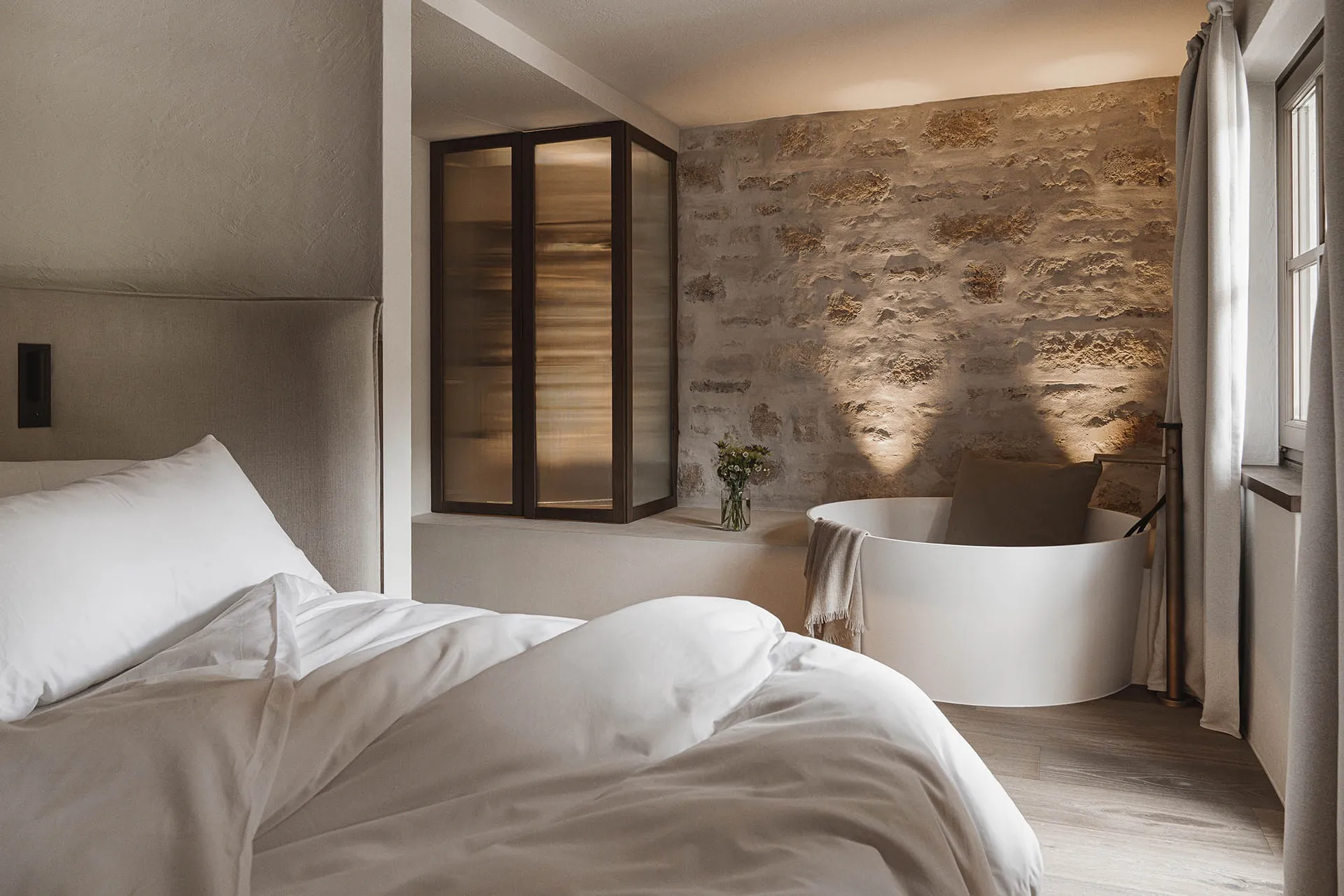 Cozy bedroom with a stone wall, a bed with a white comforter, a bathtub, and a vase with flowers on the window sill