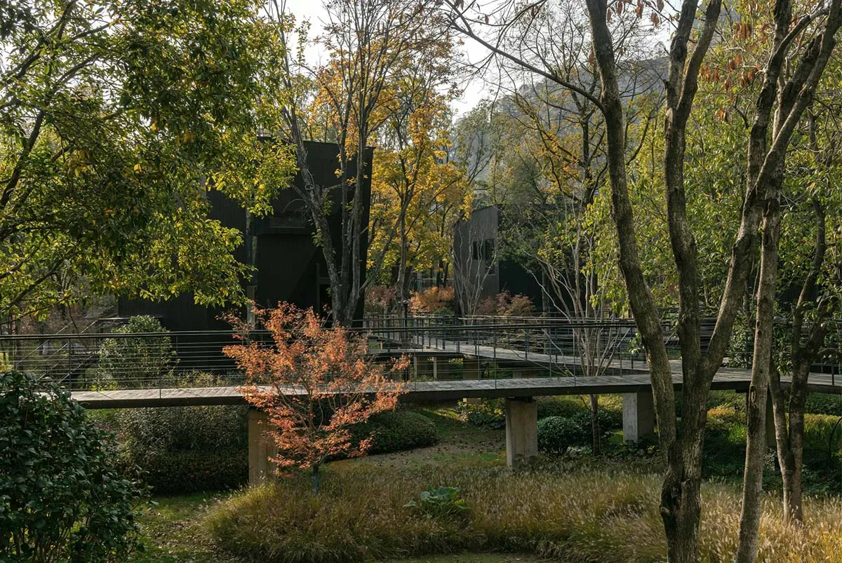 A serene outdoor space with a house surrounded by trees, a wooden bridge over a stream, and benches placed around the area