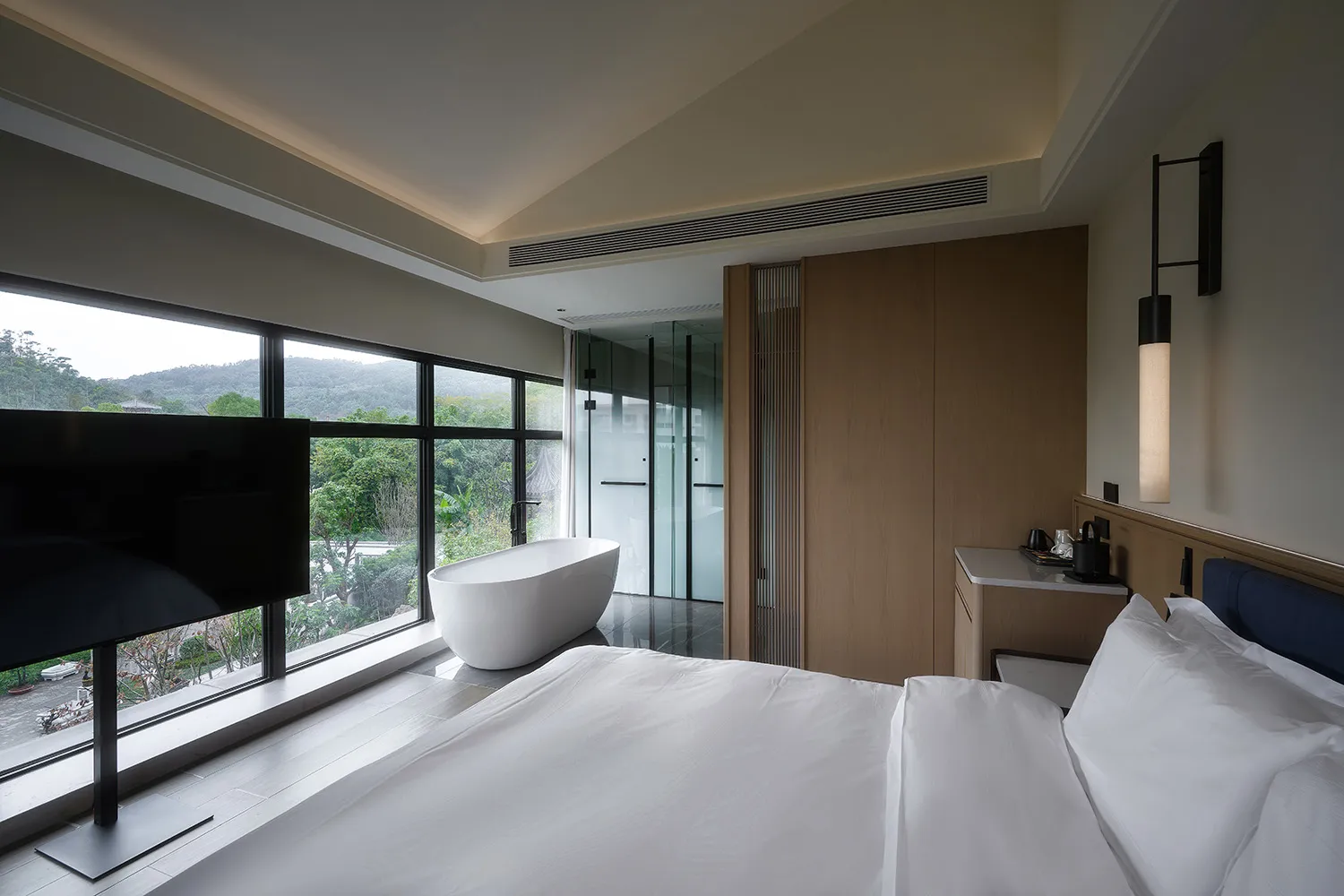 A well-lit hotel bedroom with a bed, bathtub, and wooden door, featuring a large window with a view of the outdoors
