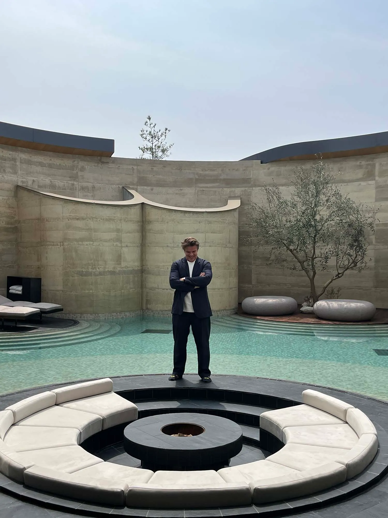 A well-dressed man poses in a large, open space with a circular centerpiece, surrounded by chairs and a potted plant, with elegant and inviting atmosphere