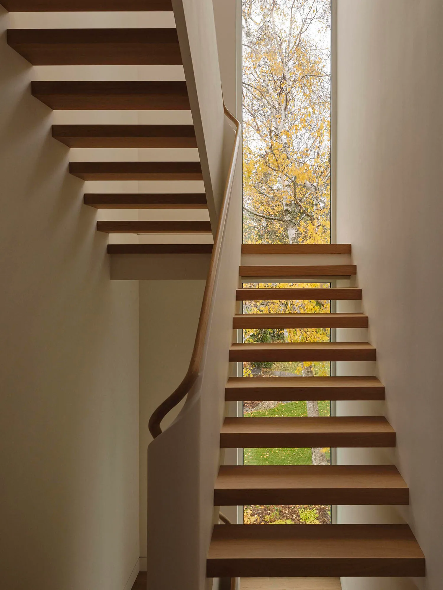 A simple staircase with a white wall and a window on the second floor, providing a view of the outside, with natural light and sunlight coming through