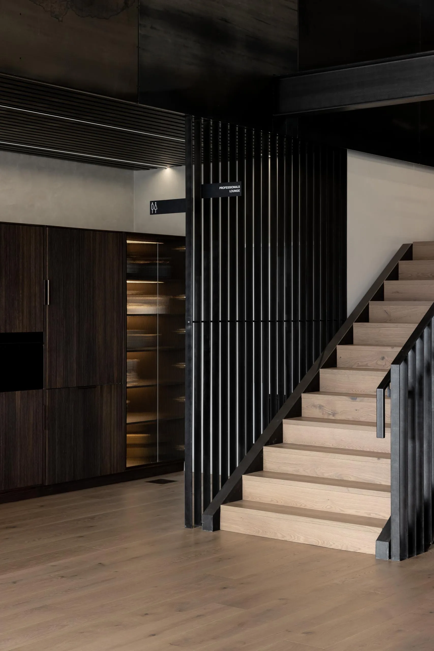 A wooden staircase with a banister in a well-lit area, possibly a living room or hallway, with a doorway at the bottom of the stairs