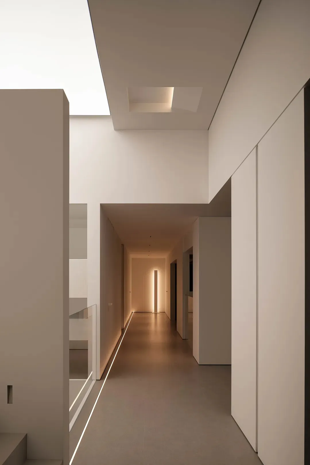 A dimly lit, narrow hallway with a white color scheme, concrete floor, and a glass door at the end