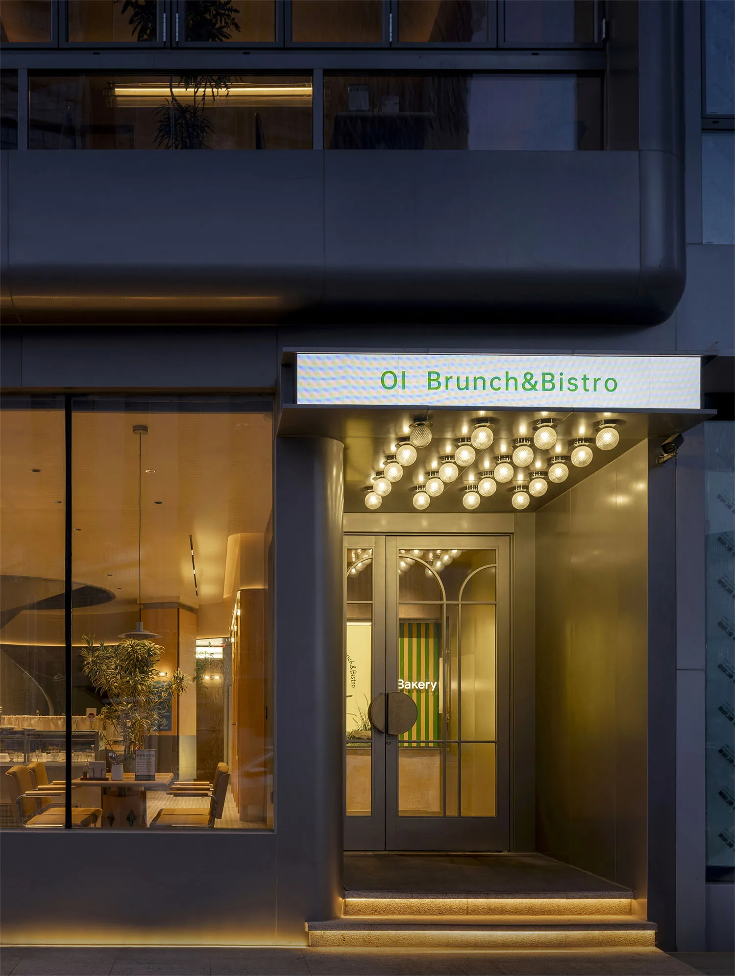 A modern restaurant entrance with a glass door, lighted sign, and warm lighting, featuring chairs and dining tables inside