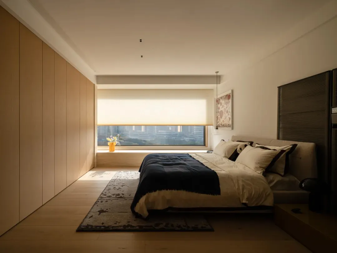 A well-lit bedroom with a bed, chair, and window, featuring wood and glass materials in a neutral color palette