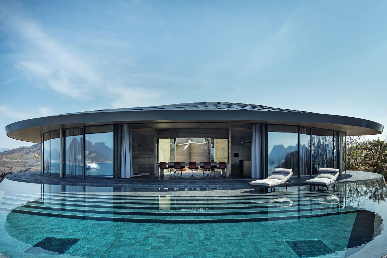 Modern open-concept living space with glass walls, a dining area, two couches, and a TV, surrounded by potted plants and overlooking a pool