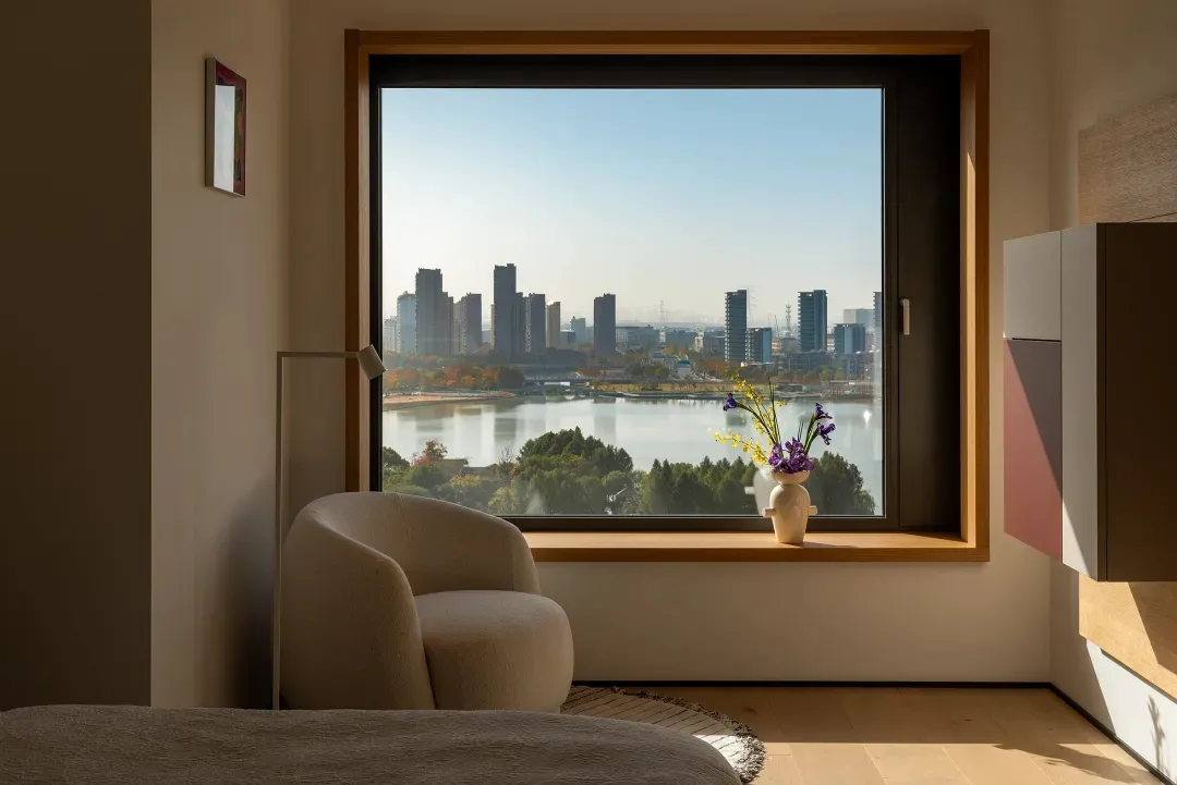 A modern living room with a couch and chair near a large window offering a city view, with a vase and flowers on a table, and natural light creating a bright atmosphere
