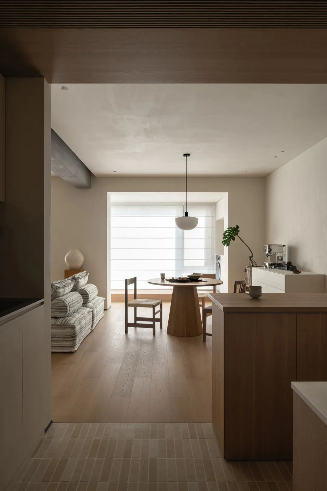 A well-lit living room and kitchen area with a couch, chair, dining table, sink, refrigerator, microwave, potted plant, and vase, featuring wood and tile materials