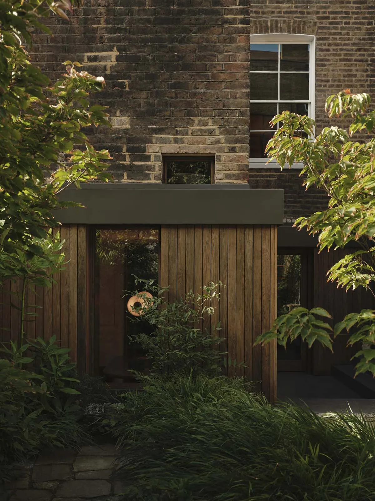 A two-story house with a brick exterior, wooden door, and window, set in a garden with a tree and bush, exuding a calm and serene atmosphere