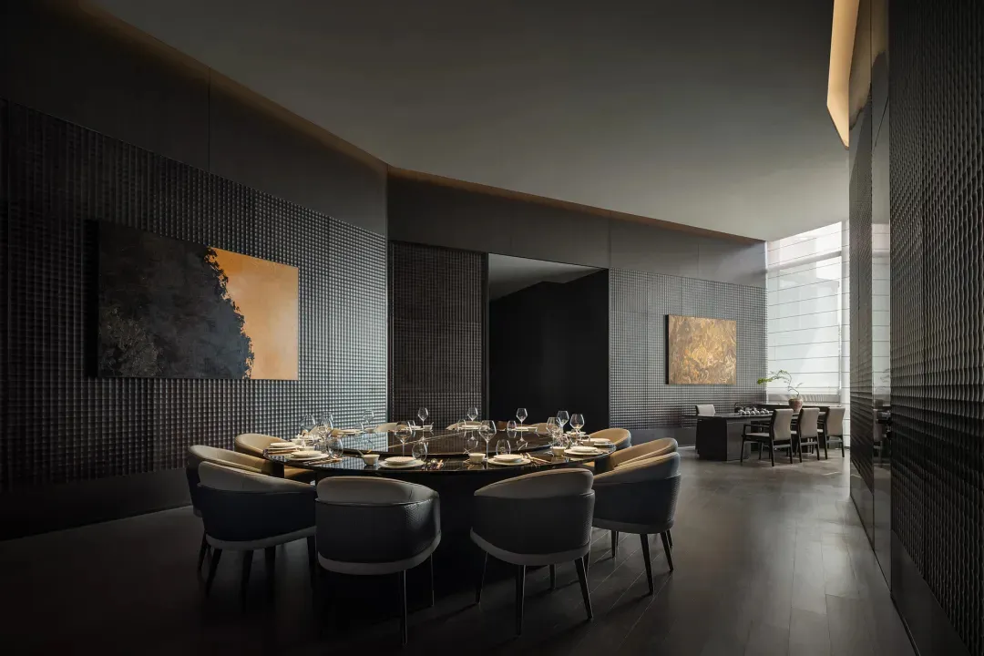 A modern dining room with a large table set with wine glasses and surrounded by chairs, featuring a couch and a potted plant in a dimly lit space with a cozy atmosphere