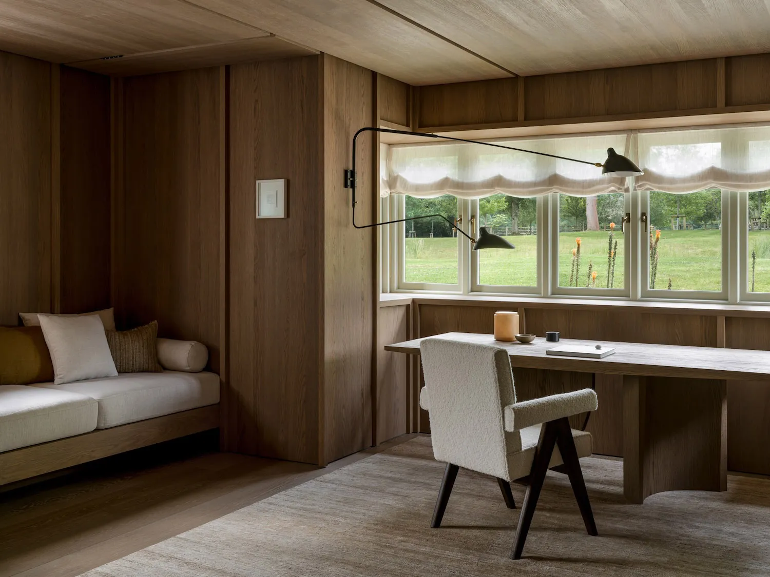 Warm and cozy living space with wooden floors and walls, featuring a couch, chair, and desk with a lamp near a window