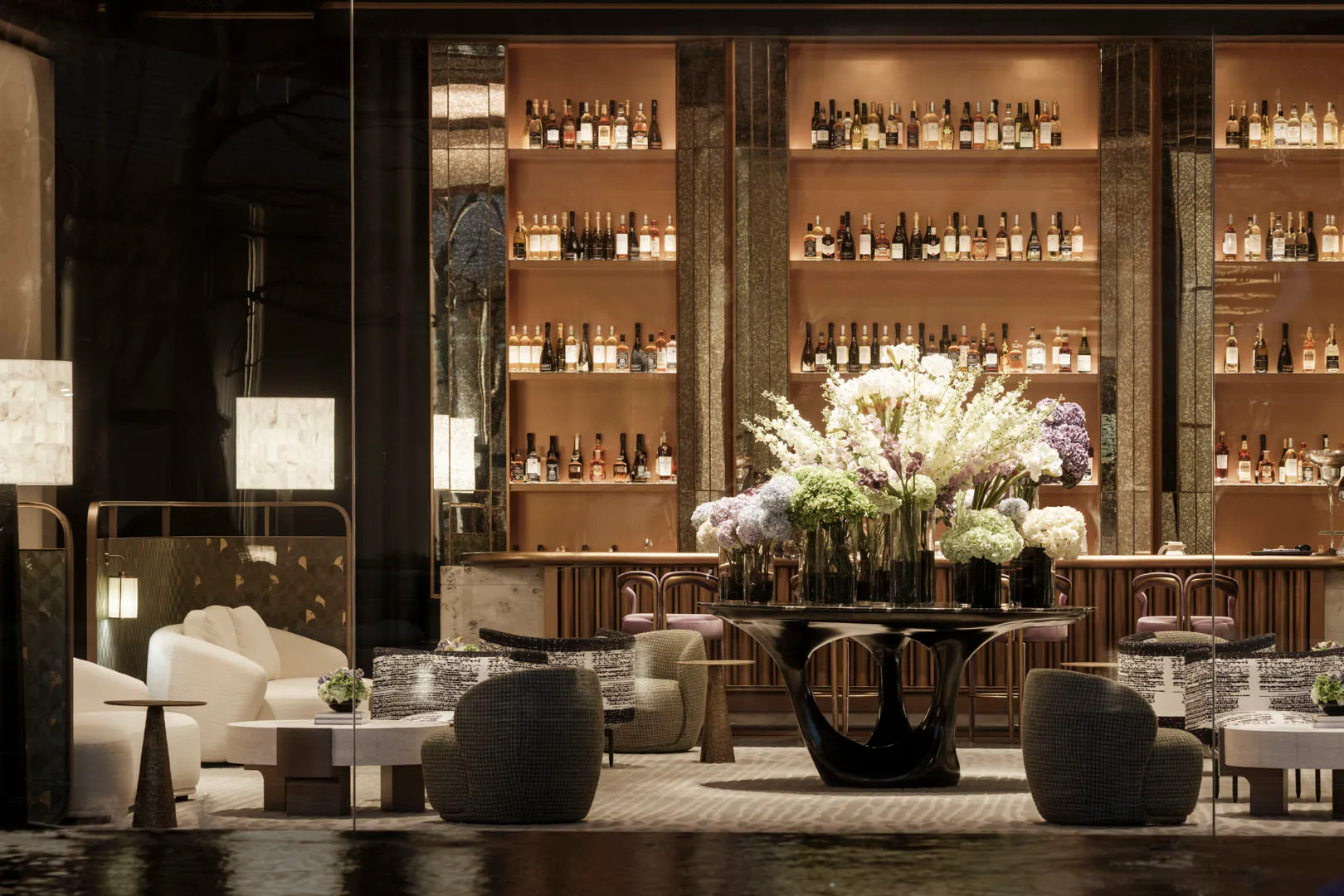 A modern bar area with a large vase of flowers, comfortable seating, and a sophisticated atmosphere, decorated with bottles and a potted plant