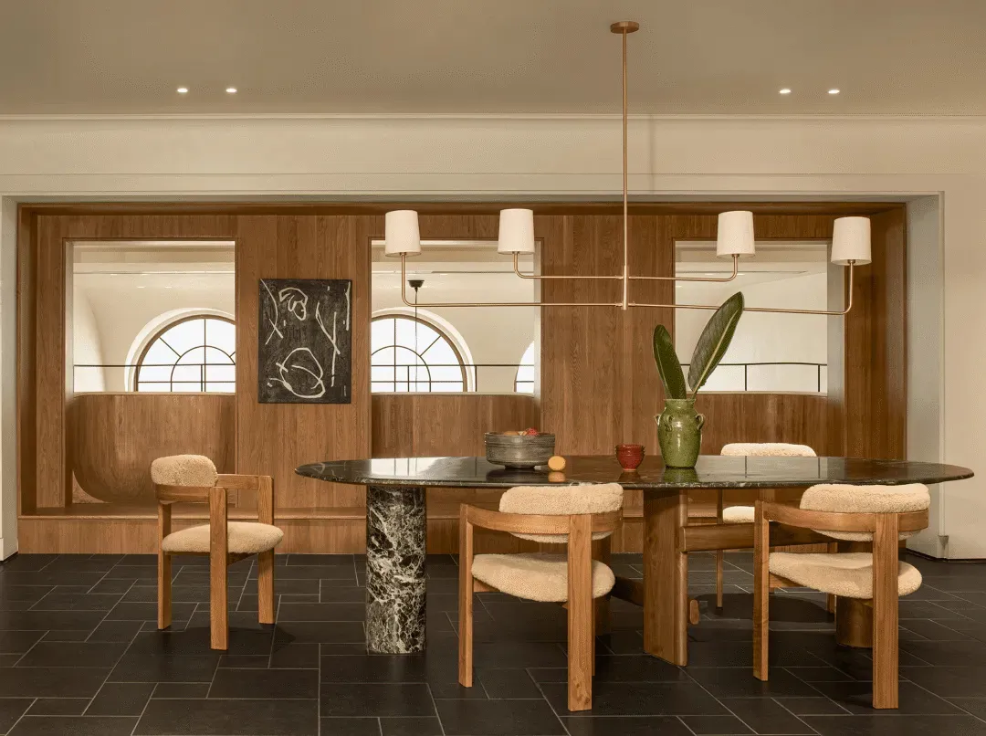 A modern dining room with a large wooden table and chairs, decorated with a bowl, vase, and potted plant, surrounded by natural light from the windows