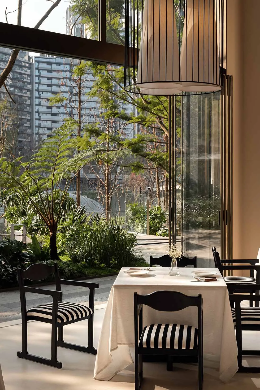 A modern dining area with a wooden table, chairs, and a lamp, set with plates, bowls, and a vase, near a window with natural light