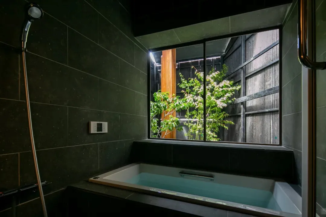 Bathroom with a black and white color scheme, featuring a bathtub with a window above it, allowing natural light to enter the room, with a tree visible outside