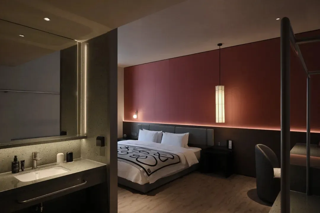 A modern bedroom with a red wall, grey countertop, a bed in the center, a chair near the right side, and a sink on the left side, with dim lighting creating a cozy atmosphere